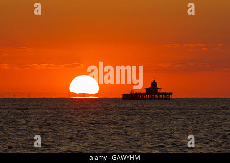 Herne Bay, Kent, Regno Unito. 6 Luglio 2016: Regno Unito Meteo. Sunrise dà il cielo di un rosso-arancione glow sopra i resti di Herne Bay pier, che era così gravemente danneggiato dalle tempeste nel 1978 che la sezione centrale è stata smantellata. Il London array, più grandi centrali eoliche offshore in tutto il mondo viene visto attraverso il calore haze. Temperature potrebbero raggiungere nella metà 20s durante il fine settimana come aria calda dal continente copre il sud del paese Credito: Alan Payton/Alamy Live News Foto Stock