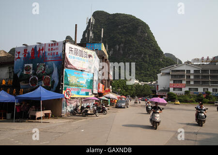 Motociclette e biciclette a cavallo su una strada e sullo sfondo si vede la zona di Guilin colline carsiche e i cartelloni in cinese. Yangshuo, nel Guangxi. Cina. Foto Stock