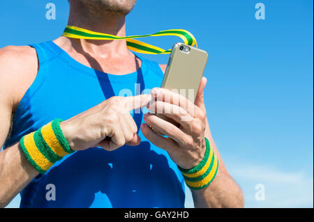 Atleta brasiliano con il giallo e il verde del Brasile di braccialetti a colori utilizzando la medaglia d'oro telefono mobile contro il cielo blu Foto Stock