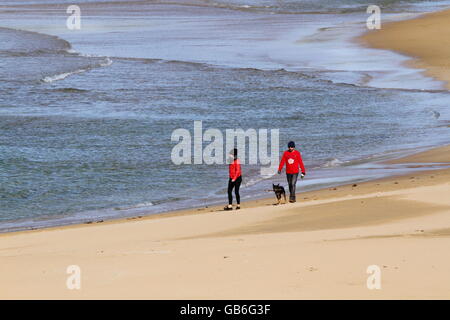 Un giovane, lady indossando giacca rossa, uomo un maglione rosso, camminare il loro cane lungo Bombo Beach, Kiama, NSW in inverno. Foto Stock