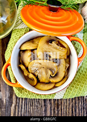 Gli champignon fritti in un rosso ceramica pentola su un tovagliolo, aglio, aneto e olio vegetale sullo sfondo dei pannelli in legno sulla parte superiore Foto Stock