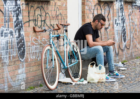 Uomo seduto sul passaggio utilizzando il telefono cellulare con la bicicletta dal lato appoggiato contro la parete con graffiti a Londra nel mese di luglio Foto Stock