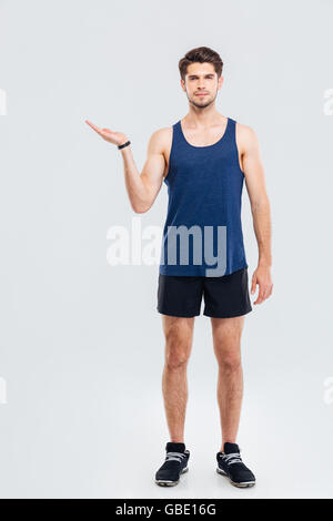 A piena lunghezza Ritratto di bello fiducioso attraente giovane atleta azienda copyspace sul palm su sfondo grigio Foto Stock
