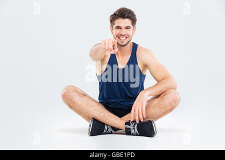 Allegro giovane atleta seduta con le gambe incrociate e puntando su di voi su sfondo bianco Foto Stock