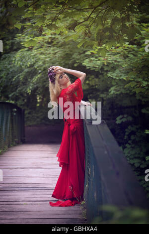 Modello bionda, delicate funzioni, viola corona di fiori, alato eyeliner, enormi ciglia, rosso Abito in pizzo, contemplativo sul ponte Foto Stock