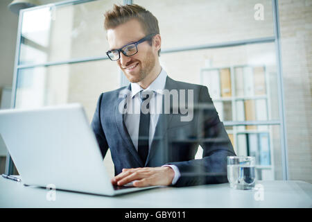 Imprenditore sorridente indossando occhiali usando il suo computer portatile al lavoro Foto Stock
