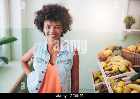 Ritratto di giovane felice juice bar proprietario guardando la fotocamera e sorridente. African modello femminile in piedi dal ripiano di frutta al cafe'. Foto Stock