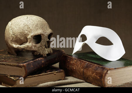 Concetto di morte. Cranio umano sul vecchio libro vicino a maschera bianca Foto Stock