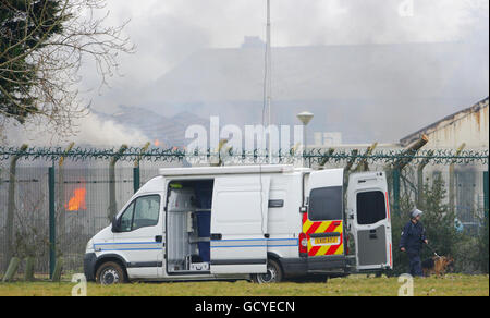 Un gestore di cani dell'ufficio di prigione specializzato cammina oltre un edificio di palestra in fiamme mentre pattugliava fuori dalla recinzione perimetrale a HMP Ford vicino Arundel, Sussex occidentale dopo circa 40 prigionieri hanno iniziato una rivolta e stabilito edifici in condizioni di luce nella prigione aperta, secondo il Ministero della Giustizia. Foto Stock