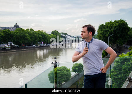 Giovane uomo bello con e-sigaretta vaping e permanente sulla terrazza vicino al fiume di città europea Foto Stock