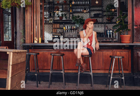 Giovane donna con cappello di fumare in un bar. Azienda di sigarette e di guardare lontano. Foto Stock
