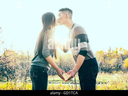 Una giovane coppia asiatica baciare in un parco in autunno e facendo un cuore con le loro mani nel tepore del sole al tramonto; Edmonton, Alberta, Canada Foto Stock