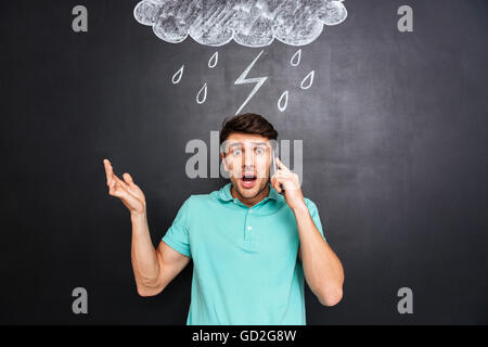 Confuso angry young man standing con telefono cellulare su sfondo blackboard Foto Stock