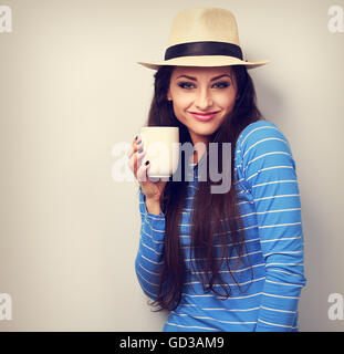 Carino felice donna informale nel cappello di paglia tenendo in mano tazza di tè e sorridente su sfondo blu Foto Stock