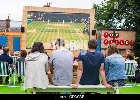 Persone prese da dietro la visione di un torneo di Wimbledon match di tennis nel grande schermo per esterni nei campi pop, parte del luogo alla moda Pop Brixton Foto Stock