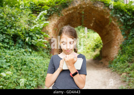 Piccola ragazza adolescente con smartwatch disegno in notebook vicino al bel vecchio ponte di arco nel parco. Carino ispanica ragazza di viaggio wri Foto Stock