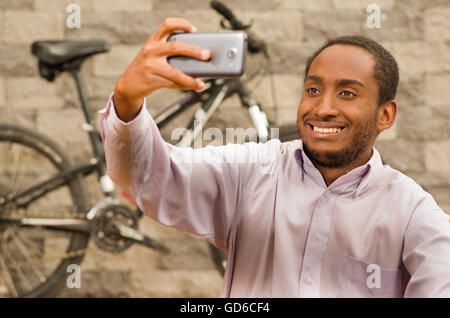 Uomo che indossa bianco rosso business shirt seduti, tenendo in mano il telefono cellulare tenendo selfie foto, sorridente e posa, noleggio in piedi dietro appoggiata contro il grigio della parete di mattoni Foto Stock