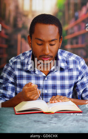 I religiosi uomo seduto mentre si tiene il Rosario, preghiera e la lettura dal libro aperto sulla scrivania davanti, il concetto di religione Foto Stock