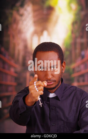 Sacerdote cattolico indossando il tradizionale collare clericale shirt in piedi di fronte alla fotocamera, tenendo le mani con rosario croce, guardando in avanti, il concetto di religione Foto Stock