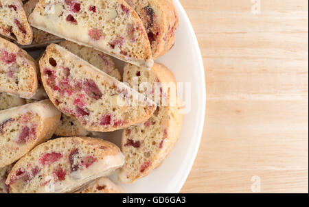 Top vista ravvicinata di biscotti con mirtilli e mandorle immersi nel cioccolato bianco su una piastra bianca in cima a una tavola di legno. Foto Stock