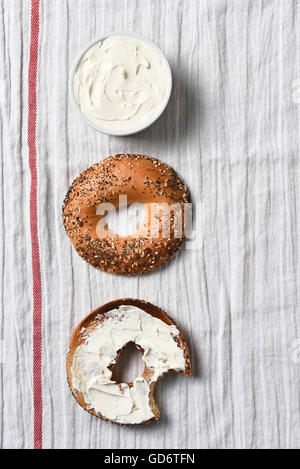 Vista aerea di un bagel affettato, una metà spalmata con la crema di formaggio. Una brocca piena di diffusione su un asciugamano da cucina. Foto Stock