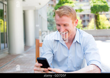 Closeup ritratto, giovani happy businessman in maglietta blu seduta, controllando il suo cellulare, isolato sullo sfondo di una città Foto Stock