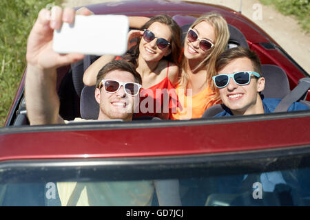 Amici guida in auto cabriolet e tenendo selfie Foto Stock