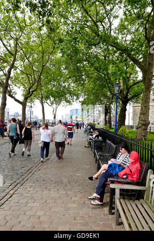 I turisti sedersi sulle panchine sotto gli alberi sul Victoria Embankment lungo il Tamigi Tower of London è sulla destra Foto Stock