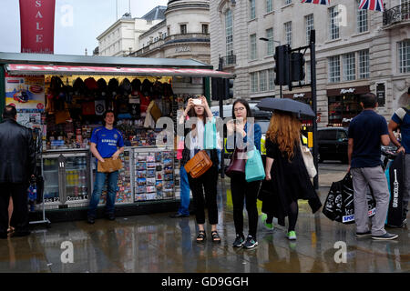 Londra Piccadilly Circus. I turisti scattano fotografie in piedi di fronte a un curio venditori stand in un giorno di pioggia. Foto Stock