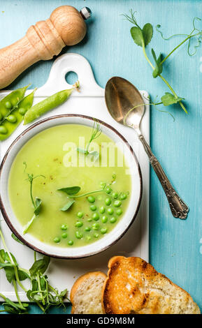 Luce estate pisello verde minestra in crema in tazza servita con i germogli, crostini di pane e spezie in ceramica bianca scheda sopra il turchese blu Foto Stock