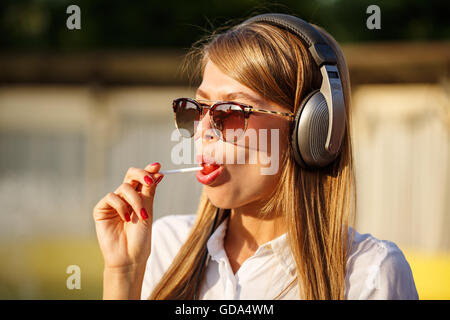 Ragazza leccare un lecca-lecca e ascolto di musica in cuffia. La ragazza di occhiali da sole Foto Stock