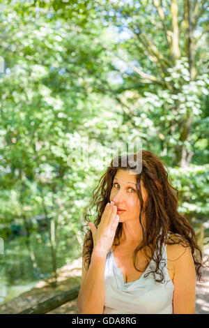 Close up busty elegante donna matura sorridente verso la telecamera mentre copre la bocca con la mano contro il giardino verde con uno sfondo con spazio di copia Foto Stock
