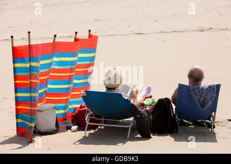 Regno Unito Galles, Gwynedd, Barmouth, spiaggia, coppia di anziani rilassarsi leggendo al sole accanto a frangivento Foto Stock