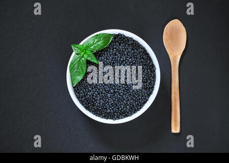 Bianco rotondo ciotola piena di lenticchie beluga con il cucchiaio di legno accanto ad essa su sfondo nero. Lay piatto con spazio di copia Foto Stock