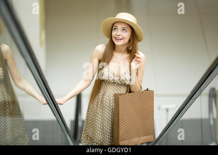 Giovane bella donna che indossa un cappello e carino polka dot dress portando shopping bag stando in piedi sulla scala mobile nel centro commerciale Foto Stock