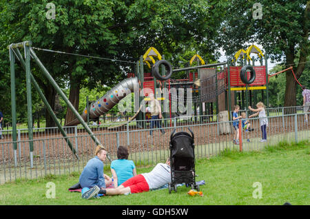I genitori rilassarsi sul prato mentre i bambini giocano nel parco giochi a Prospect Park, la lettura. Foto Stock