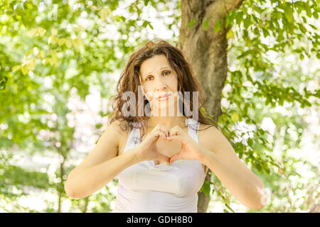 Bella donna fare a forma di cuore con le mani contro il giardino verde sullo sfondo Foto Stock