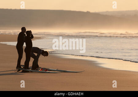 Tre surfisti in silhouette con tavole da surf permanente sulla spiaggia al tramonto guardando il set di onda Foto Stock