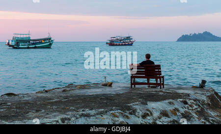 Un solitario giovane uomo seduto sul molo sul mare al tramonto. Foto Stock