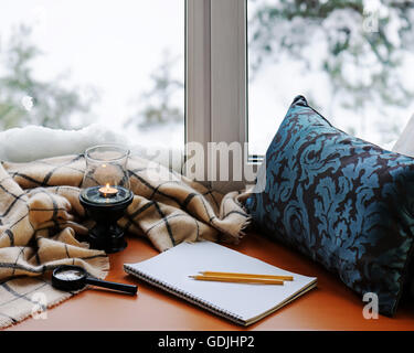 Aprire il blocco note, vetro magnifier, cuscino, candela, matite e beige caldo plaid situato sul legno stilizzata davanzale. Inverno concep Foto Stock