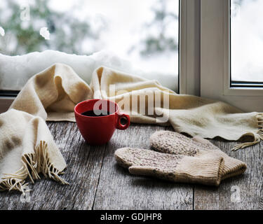 Red tazza di caffè o di tè, beige sciarpa e womans guanti sono situati sul legno stilizzata davanzale. Inverno concetto di comfort di un Foto Stock