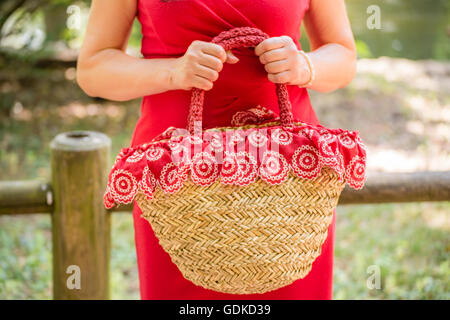 Mani femminili tenendo un sacco in un paese stile fatto di rafia e panno rosso con fiori di colore bianco, la donna vestita in una guaina rossa vestito è in attesa nervosamente in un parco Foto Stock