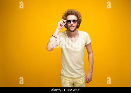 Ritratto di curly-pelose giovane uomo in occhiali da sole e bermuda cercando confuso a telecamera. Isolare. Sfondo giallo. Foto Stock