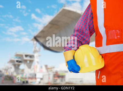 Protezione di sicurezza delle attrezzature di lavoro. Lavoratore in arancione vest azienda Casco giallo contro il sito in costruzione come sfondo Foto Stock