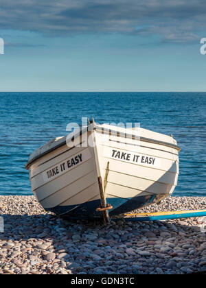 Take it easy - Mare Scena raffigurante un oceano blu di sfondo con barca a metà del terreno e una spiaggia di ciottoli in primo piano. Foto Stock