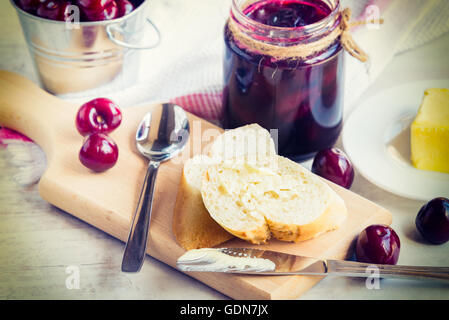 Toast imburrato e un vaso con cherry marmellata fatta in casa bianco su uno sfondo di legno Foto Stock