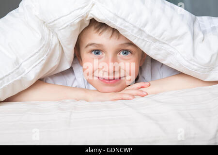 Ragazzo sorridente nascondendo nel letto sotto una coltre bianca o coverlet. Foto Stock