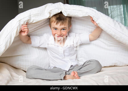 Ragazzo sorridente nascondendo nel letto sotto una coltre bianca o coverlet. Foto Stock