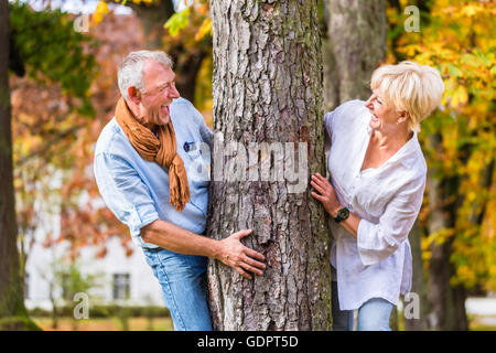 Coppia senior, uomo e donna, flirtare con ogni altro di giocare a nascondino intorno a un albero in albero di caduta Foto Stock