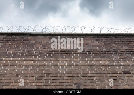 Guardando verso l'alto un alto muro di mattoni con filo spinato sulla parte superiore e le nuvole scure sopra Foto Stock